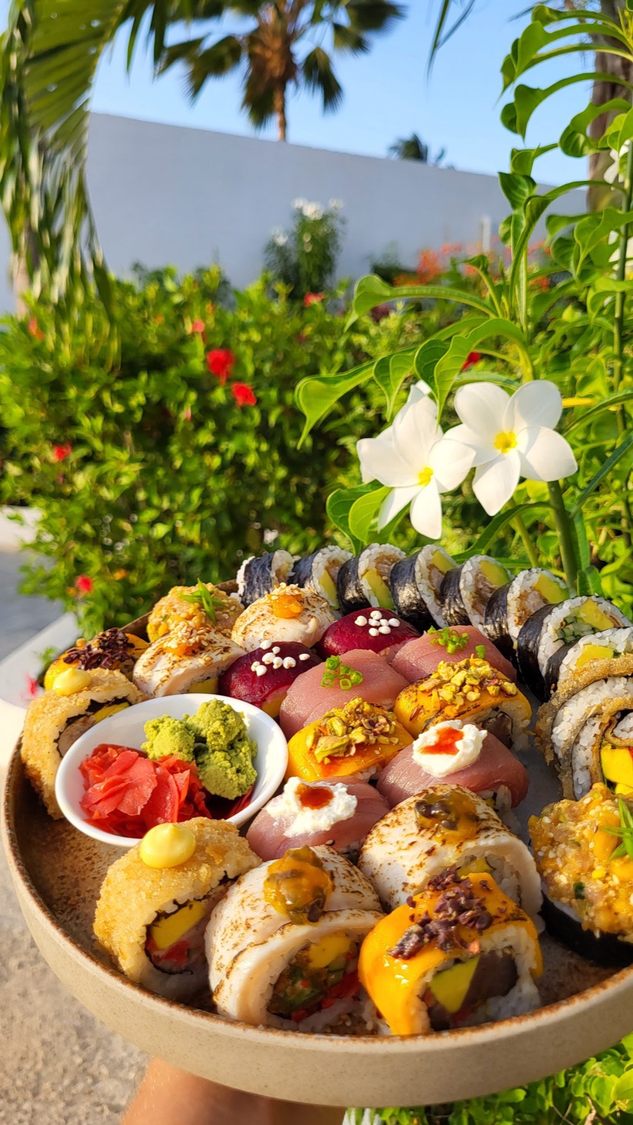 A generous KAMI sushi platter held up against Zanzibar palms and frangipani flowers